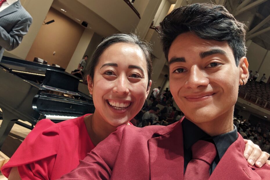 Dr. Jessica Lu, dressed in red, appears on the left with Bryant Rivera Cortex, dressed in a black shirt, maroon jacket and tie. A black piano is in the immediate background, with the members of the Honors College Class of 2026 departing the concert hall seats in the further background.